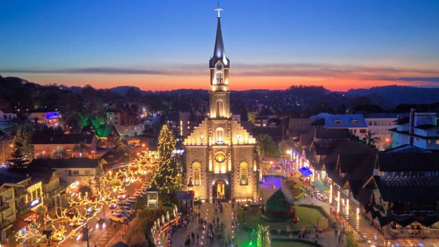 Catedral na cidade de Gramado, no Rio Grande do Sul, ao entardecer, com o pôr-do-sol ao fundo e com as ruas iluminadas, com uma pequena multidão à frente do local