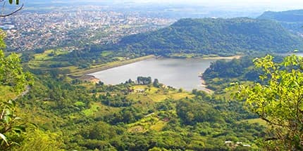 Vista aérea de Santa Maria (RS) a partir de um morro, com um lago cercado por mata verde em primeiro plano e a cidade ao fundo, sob céu claro.
